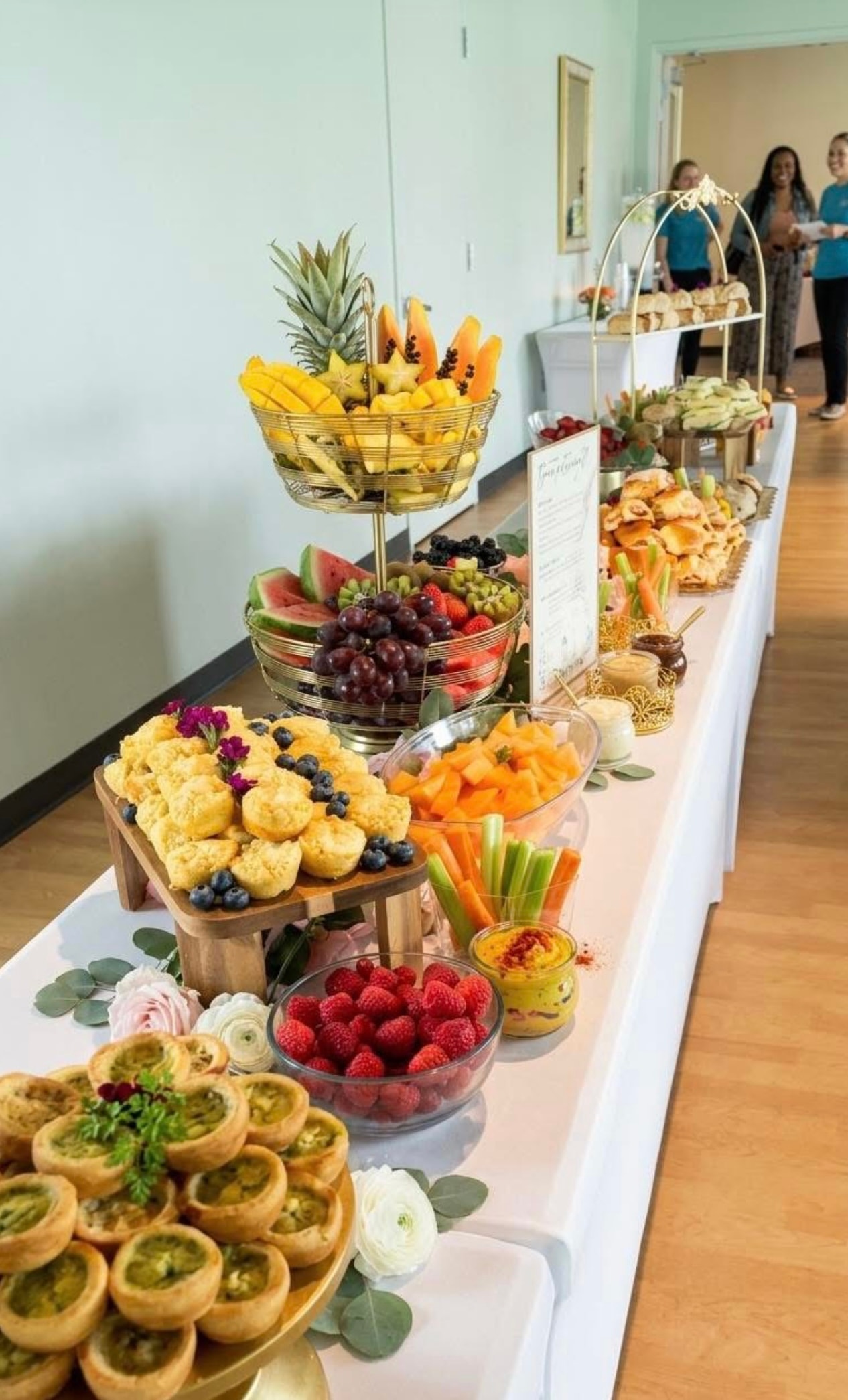 Long elegant buffet table with fruit tower, quiche cups, and florals